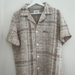 Beige kortärmad skjorta från Nikben - En perfekt kombination av stil och komfort. Denna skönhet har luftiga korta ärmar och en praktisk knäppning med pärlknappar. Med sitt avslappnade snitt passar den både herrar och damer, vilket gör den till ett utmärkt val för alla tillfällen. Oanvänd och i nyskick. Säljer då den är för liten. Nypris: 1200kr