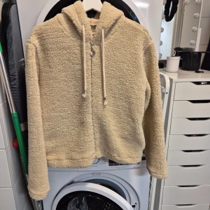 Beige teddyjacka med huva - Mysig beige teddyjacka med huva och dragkedja framtill. Perfekt för kyliga dagar när du vill hålla dig varm och bekväm. Jackan har långa ärmar och dragsko vid huvan för justerbar passform och även fickor på sidorna