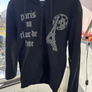 Svart hoodie med Stenar - Snygg svart hoodie från Bling lucid med dragkedja och en cool design av Eiffeltornet i glittriga stenar. Texten 'Paris au clair de lune' pryder framsidan. Perfekt för en avslappnad stil med en touch av Paris. Hör av dig vid frågor eller funderingar✅