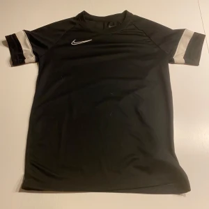 Svart fotbollströja från Nike - Snygg svart fotbollströja från Nike med korta ärmar och vita detaljer. Perfekt för träning eller match. Tillverkad i Dri-FIT-material för optimal komfort och andningsförmåga.