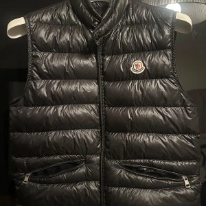 Monclear GUI väst - Snygg svart dunväst från Moncler med dragkedja och två praktiska fickor framtill. Västen har en klassisk design med Moncler-loggan.                        Skriv om du vill ha fler bilder på den eller undrar något!