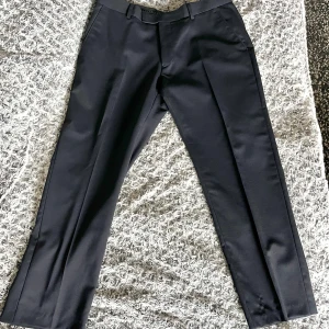 Svarta kostymbyxor - Snygga svarta kostymbyxor från det exclusive märket Riley med en klassisk rak passform. De har en midwaist design och är perfekta för en stilren look. Byxorna har pressveck där fram och är 60% ull.