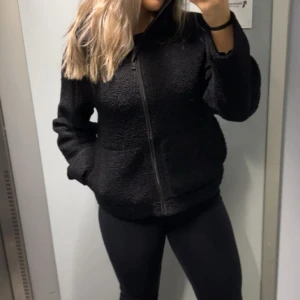 Svart teddyjacka med dragkedja - Mysig svart teddyjacka med dragkedja framtill. Perfekt för kyliga dagar med sin fluffiga och varma design. Jackan har långa ärmar och en bekväm passform som gör den idealisk för vardagsbruk.
