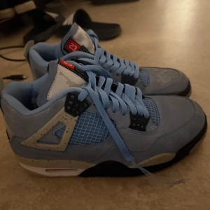 Jordan 4 University blue - Snygga Nike Air Jordans i ljusblå mocka med svarta och grå detaljer. Skorna har en klassisk snörning och den ikoniska Jumpman-loggan på hälen. Perfekta för dig som vill ha en stilren och sportig look.