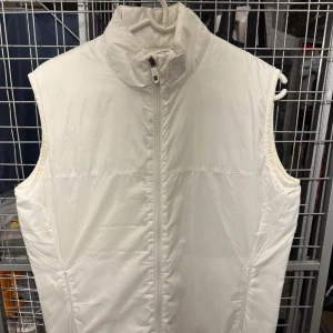 Vit dunväst med dragkedja från FootJoy - Snygg vit dunväst med dragkedja framtill. Perfekt för kyligare dagar och enkel att matcha med andra plagg. Västen har en hög krage och en stilren design.