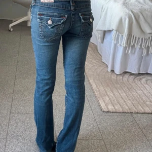 Blå jeans med broderade fickor - Snygga blå jeans från True religion storlek 25w med broderade detaljer på bakfickorna och en klassisk femficksdesign. De har en låg midja och en bootcut-stil som ger en avslappnad look. Perfekta för en casual outfit.