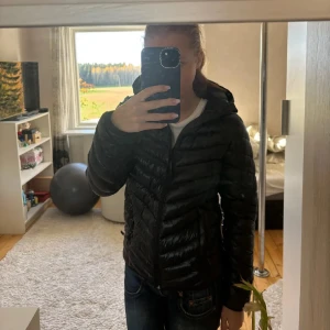 Svart dunjacka med huva - Säljer en stilren svart dunjacka med huva. Jackan har en dragkedja framtill och är perfekt för kyligare dagar. Den är lätt och smidig att bära, vilket gör den till ett utmärkt val för höst och vinter. Aldrig andvend helt ny. 