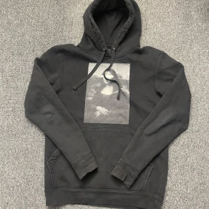 Hoodie - Cool svart hoodie med ett stort Mona Lisa-tryck på framsidan. Den har en klassisk känguruficka och justerbar huva med snören. Perfekt för en avslappnad stil.