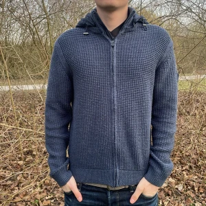 Blå stickad cardigan från Polo Club - Säljer en snygg blå stickad cardigan från Polo Club. Tröjan har en dragkedja framtill och en quiltad huva. Perfekt för kyliga dagar med sin stilrena design och bekväma passform. Lite nopprig 