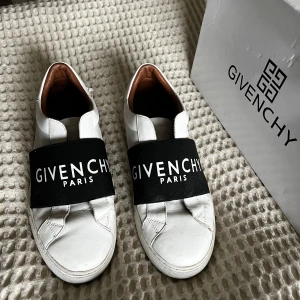 Vita sneakers från Givenchy - Snygga vita sneakers från Givenchy med en bred svart rem över foten där Givenchy Paris är tryckt i vitt. Skorna har en stilren design med rund tå och är tillverkade i skinn. Kom med bud 