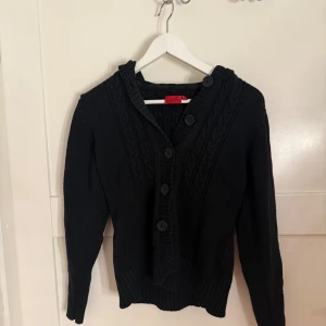 Svart stickad kofta med knappar - Snygg svart stickad kofta från Fransa med stora knappar och huva. Koftan har långa ärmar och ribbade detaljer vid ärmslut och nederkant. Storlek M men skulle säga att den är en  Xs- S. 