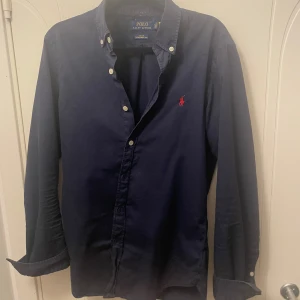 Mörkblå skjorta från Ralph Lauren - Snygg mörkblå skjorta från Ralph Lauren i slim fit. Skjortan har långa ärmar och är tillverkad i ett lätt twillmaterial perfekt till varmare dagar! Köpt förra året för 1400 men säljer för den har blivit för liten. Hör av dig vid intresse eller funderingar!