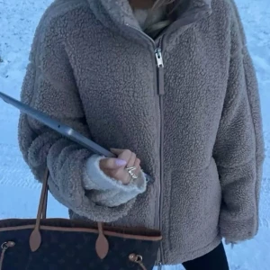 Beige teddyjacka med dragkedja - Mysig beige teddyjacka med dragkedja framtill. Perfekt för kyliga dagar med sin fluffiga och varma design. Jackan har långa ärmar och en hög krage för extra värme.