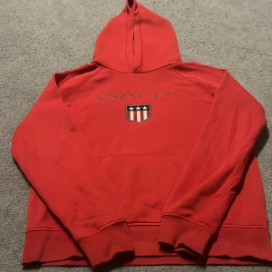 Röd hoodie från GANT - Säljer en snygg röd hoodie från GANT med ett tryckt emblem på framsidan. Tröjan har en klassisk känguruficka och en bekväm huva. Perfekt för en avslappnad stil.