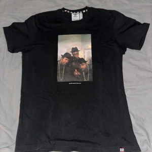 Svart t-shirt med Run DMC-tryck från Adidas - Säljer en svart t-shirt från Adidas med ett coolt Run DMC-tryck på framsidan. T-shirten har korta ärmar och är gjord i bomull. Perfekt för dig som gillar klassisk hiphop-stil. Texten 'My Adidas' finns tryckt på baksidan.