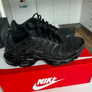 Svarta Nike Air Max Plus sneakers - Säljer ett par helsvarta Nike Air Max Plus sneakers med ikonisk vågig design och synliga Air-enheter i sulan. Ovandelen är i mesh och syntet med snygga detaljer och klassisk Nike-logga på tungan. Grymma skor för dig som gillar streetstyle och komfort.