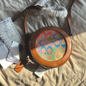 Coach rund axelväska med färgglatt mönster - Unik väska!! Cool. Kunde inte låta bli. Säljer en rund axelväska från Coach med bruna skinnkanter och ett färgglatt mönster med Coach-loggor i blått, rosa, grönt, lila och gult. Väskan har justerbar axelrem i brunt skinn och guldfärgad logga framtill. Perfekt för dig som vill sticka ut med en unik accessoar.inköp 6800. 