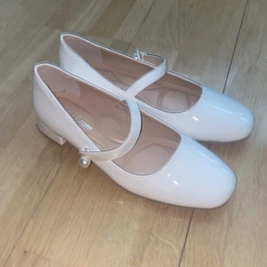 Vita Mary Jane skor från Abloom - Snygga, oanvända, vita Mary Jane ballerinaskor från Abloom med lackad finish, låg klack och rund tå. Remmen har en pärldetalj som ger en extra touch. Perfekta för dig som gillar klassisk och söt stil. storlek 37 på skon men skulle nog säga att det är storlek 38. 