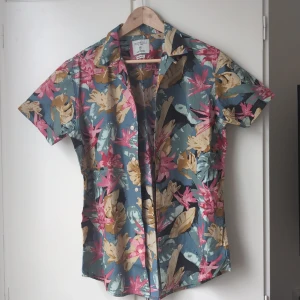 Blommig kortärmad skjorta Springfield M - Färgglad kortärmad skjorta från Springfield i storlek M. Skjortan har ett blommigt mönster i rosa, beige och blått med knappar framtill och klassisk krage. Perfekt för dig som vill sticka ut med en somrig vibe.