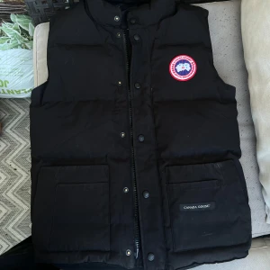 Svart dunväst från Canada Goose - Svart dunväst från Canada Goose med klassisk logga på bröstet och två stora fickor framtill. Västen har hög krage, dragkedja och tryckknappar. Perfekt för lager-på-lager och håller dig varm under kyliga dagar.
