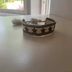 Flätat armband  - Snyggt vävt armband i beige med svarta stjärnor runt om. Armbandet har knytning och två dekorativa tofsar i ändarna. Perfekt accessoar för att ge din outfit en cool touch. Skriv till mig vid minsta lilla fundering!