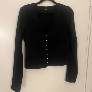 Svart v-ringad kofta med pärlknappar - Säljer en svart v-ringad kofta med långa ärmar och snygga pärlknappar framtill. Perfekt att styla till olika outfits för en stilren look.