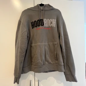 Grå hoodie från Hoodrich - !! Pris kan diskuteras !!Säljer en grå hoodie från Hoodrich med broderad logga i svart och vitt på bröstet samt röd text under. Tröjan har huva med dragsko och en stor magficka. Perfekt för en avslappnad stil.
