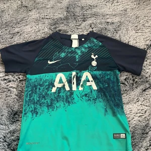 Tottenham Hotspur bortatröja Nike - Snygg Tottenham Hotspur bortatröja från Nike med korta ärmar. Tröjan har mörkblå axlar och ärmar, samt turkos och blått mönster framtill. Tryckt klubbmärke och Nike-logga på bröstet, samt AIA sponsortryck. Tillverkad i lätt och ventilerande material.