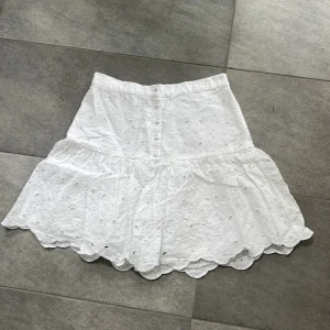 Vit broderad kjol Holly & Whyte - Superfin vit kortkjol från Holly & Whyte med broderade blommor och hålmönster. Kjolen har volangkant nertill och elastisk midja för skön passform. Perfekt för sommarens alla tillfällen och ger en fräsch och söt look.