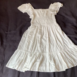 Vit sommarklänning från KappAhl - Säljer en söt vit klänning från KappAhl med korta puffärmar och smock upptill. Klänningen har volang nertill och är tillverkad i ett lätt bomullstyg, perfekt för varma dagar. Passar dig som gillar romantisk och luftig stil.