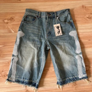 Jorts KIJITTY - Unika jeansshorts från KIJITTY med råa, fransiga benslut och coolt vitt benmönster längs sidorna. Klassisk blå denim med fem fickor och normal passform. Perfekta för dig som vill sticka ut med en edgy streetstyle.