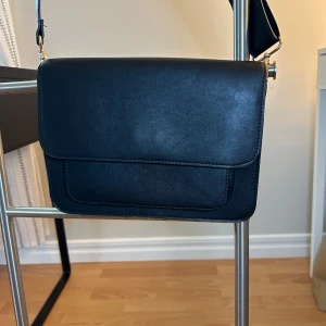 Svart axelväska i skinnimitation - Stilren svart axelväska med bred justerbar rem och magnetlås. Väskan har ett enkelt, minimalistiskt uttryck och är tillverkad i slät skinnimitation. Framsidan har en praktisk ficka och insidan är fodrad i svart tyg.