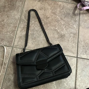 Svart axelväska med nitar och kedja - Snygg svart axelväska i skinnimitation med coolt geometriskt mönster av små silverfärgade nitar. Väskan har en kedjerem och ett stort fack med dragkedja inuti. Perfekt för dig som vill ha en edgy och trendig look.