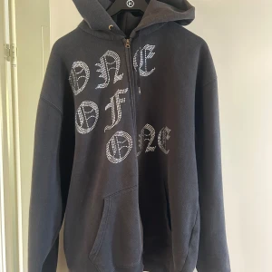 Svart hoodie från One of One - Säljer en svart hoodie från One of One i storlek medium. Tröjan har dragkedja, huva med snören och stora vita bokstäver med stenar på framsidan. Perfekt för dig till sommaren.