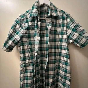 Grön och vit rutig kortärmad skjorta - Snygg kortärmad skjorta i grönt och vitt rutigt mönster. Skjortan har klassisk krage, knappar framtill och är tillverkad i mjuk bomull. Perfekt för en avslappnad och cool stil.