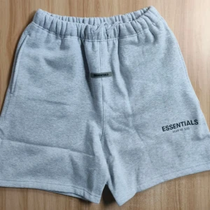 Grå Essentials shorts med snörning - Ljusgrå shorts från Essentials Fear of God med elastisk midja och snörning framtill. Mjukt bomullsmaterial, två sidofickor och en bakficka. Diskret logga på vänster ben. Perfekta för chill eller träning.