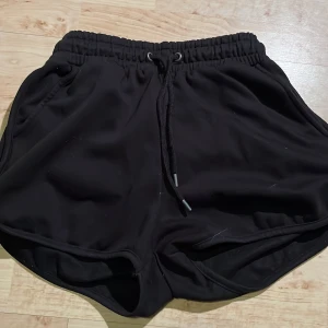 Svarta träningsshorts från Eltzia - Svarta träningsshorts från Eltzia med elastisk midja och snörning. Modellen är kort och har en sportig look med rundade benslut och fickor på sidorna. Perfekta för gymmet eller löprundan.