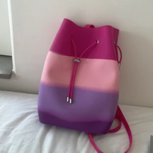 Färgglad ryggsäck med bläckfiskberlock - Säljer en unik ryggsäck i rosa, lila och ljusrosa med cool snörning upptill och justerbara axelband. Väskan har en silvrig bläckfiskberlock som detalj och är tillverkad i ett syntetmaterial som känns mjukt men ändå stabilt. Perfekt för dig som vill sticka ut med färg och lekfull stil.