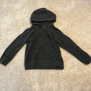Mörkgrå stickad hoodie från JJXX - Mörkgrå stickad hoodie från JJXX med långa ärmar och huva. Perfekt för kyliga kvällar och enkel att matcha med olika outfits. Klassisk modell med ribbade muddar vid ärmslut och nederkant. Endast testad.