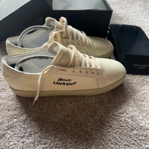 Vita sneakers från Saint Laurent - Säljer ett par stilrena vita sneakers från Saint Laurent med svart logga på sidan och diskret branding på häl och plös. Skorna har snörning och är tillverkade i canvas och skinn. Perfekta för dig som gillar minimalistisk design.