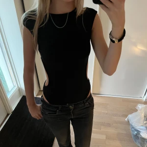 Svart body med cut-outs - Säljer en svart body med tight passform och coola cut-outs på sidorna. Toppen är ärmlös och har en rund halsringning. Perfekt för dig som vill sticka ut lite extra!