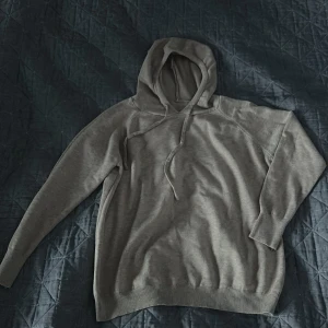 Grå hoodie med huva - Säljer en enkel och stilren beige hoodie med huva och dragsnören. Tröjan har långa ärmar och ribbade muddar vid ärmslut och nederkant. Perfekt för en avslappnad look.