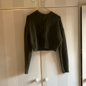 Mörkgrön croppad kofta - Säljer en mörkgrön croppad kofta med rund halsringning och knappar framtill. Tröjan är stickad och har långa ärmar. Perfekt att styla med höga jeans eller kjol.