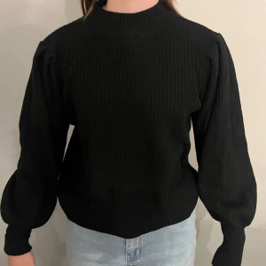 Svart ribbad stickad tröja med puffärmar - Säljer en svart stickad tröja med ribbad struktur och trendiga puffärmar. Tröjan har hög hals och lång ärm, perfekt för att lyfta din outfit. Snygg till både jeans och kjol!