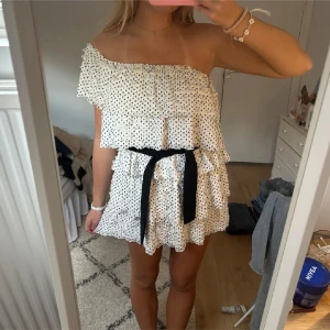 Vit prickig omlottklänning med volanger - Går att ha utan band med båda axlar, utan axlar och med en axel perfekt!Säljer en söt vit kortklänning med svarta prickar och volanger. Klänningen är oneshoulder och har ett svart knytband i midjan. Perfekt för sommarens alla tillfällen!