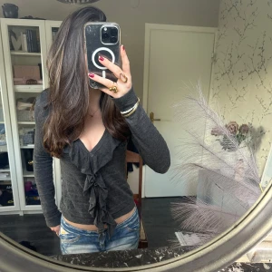 Mörkgrå kofta med volang - Säljer en mörkgrå långärmad kofta med knappar och v-ringning och volangdetaljer framtill. Koftan har knyt i midjan och är i ett mjukt, stretchigt material. Perfekt för dig som gillar trendiga och feminina plagg. Har lite klittertrådar i materialet. I storlek xs-s