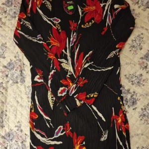 Svart långklänning med rött blommönster från NVF Fashion Paris - Helt ny. Säljer en svart långklänning med färgstarka röda och vita blommor från NVF Fashion Paris. Klänningen har lång ärm och hög hals, samt ett snyggt veckat tyg som ger struktur. Perfekt för dig som vill sticka ut med ett unikt mönster. 