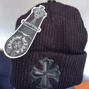 Svart ribbad mössa från Chrome Hearts - Svart ribbstickad mössa från Chrome Hearts med ett stort svart kors i skinn framtill. Klassisk design med uppvikt kant och tydlig logga på etiketten. Perfekt accessoar för en edgy streetwear-look.