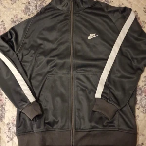 Grå träningsjacka från Nike - Säljer en grå träningsjacka från Nike med vita ränder längs ärmarna och dragkedja framtill. Jackan har ribbade muddar och klassisk Nike-logga på bröstet. Perfekt för sportiga tillfällen eller chill vardagsstil.