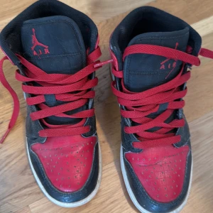 Nike Air Jordan 1 Mid Black/Red - Säljer ett par klassiska Nike Air Jordan 1 Mid i svart och rött med vita sulor. Skorna har röd snörning, svart läder och röda detaljer samt Air Jordan-logga på sidan. Perfekta för dig som gillar streetwear och sneakers med ikonisk design.